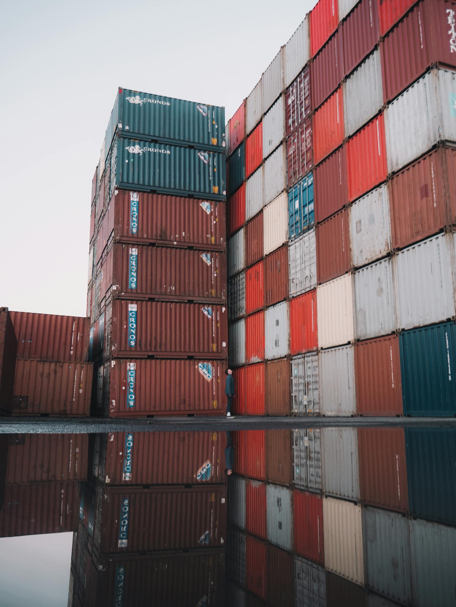 Stacks of colorful cargo containers at an industrial shipping yard, reflecting on wet ground.